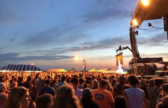 A large crowd at an outdoor concert during twilight, with a performer on stage, several tents in the background, and stage lights illuminating the scene.