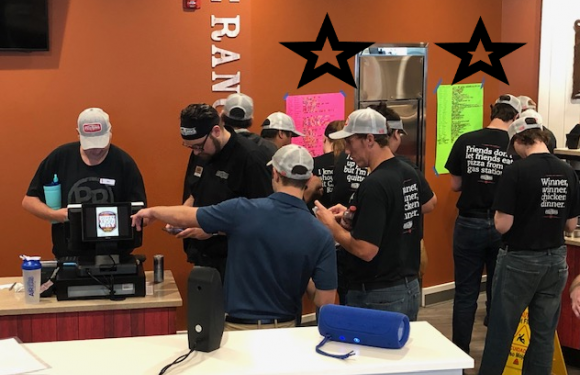 Employees in black and navy uniforms and gray caps work together behind a counter at Pizza Ranch, with colorful menus on an orange wall in the background.