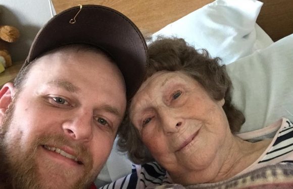 A young man in a red shirt and baseball cap taking a selfie with an elderly woman lying in a bed, wrapped in a patterned blanket, in a room with a wooden headboard.