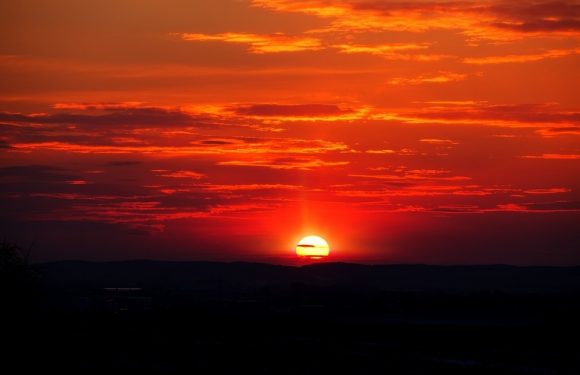 A vibrant sunset with the sun partially below the horizon, casting orange and red hues across a sky filled with layered clouds over a dark landscape.