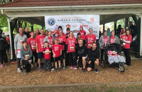 Group of people wearing red shirts, some with race bibs, standing under a gazebo with a banner for the Orange City Tulip Festival and Kiwanis Road Race.