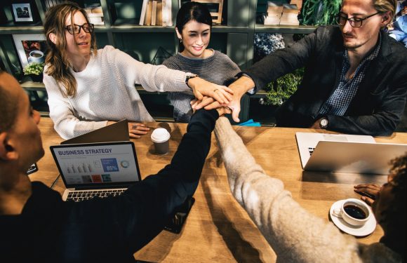 Six diverse people sit around a wooden table with laptops and coffee, placing their hands together in a gesture of teamwork and unity.