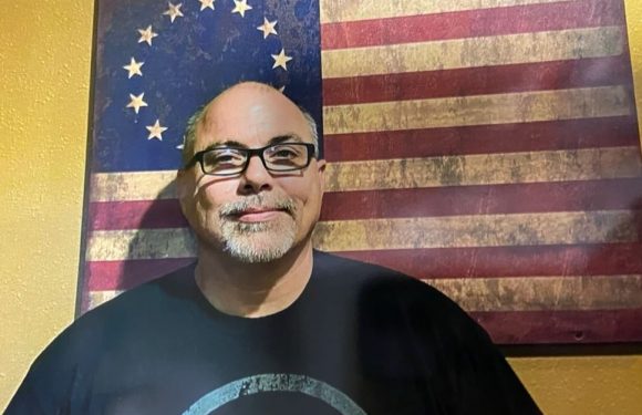 A man with glasses, a goatee, and a mustache is smiling in front of a rustic American flag hanging on a yellow wall.