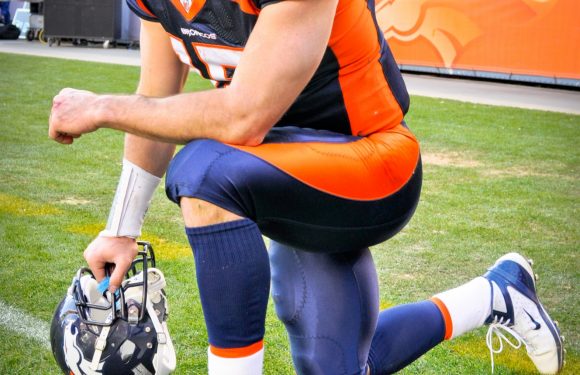 A football player in a blue and orange uniform kneels on the field, holding his helmet, with a crowd in the stadium background.