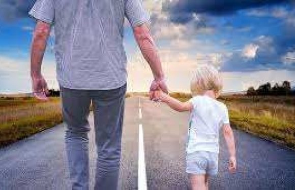 A person and a child holding hands and walking down an open road with grass fields on either side, under a partly cloudy sky.