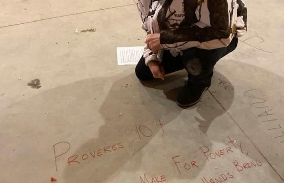 A man wearing a Pizza Ranch cap and a patterned jacket crouches on a concrete floor, next to a handwritten sign with a quote about wealth and effort.