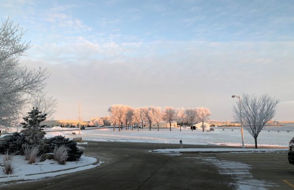 Snow-covered landscape with leafless trees, a parking lot, and a few buildings in the distance under a partly cloudy sky during winter.