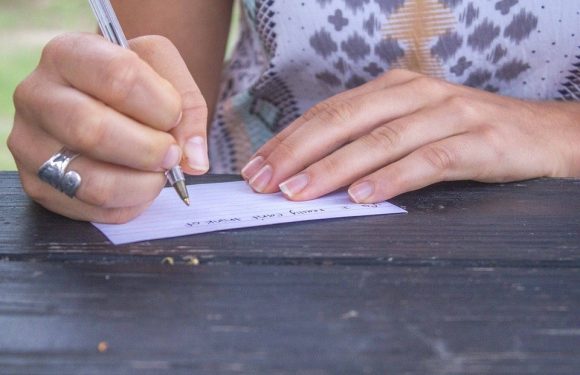 A person writes a note on lined paper with a pen, resting their other hand on a dark wooden surface. The person wears a patterned shirt and a silver ring.