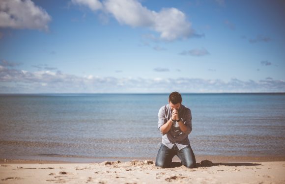 A man in a gray shirt and jeans kneels on the sandy beach near the water, holding his hands together in front of his face in a prayer or reflective pose.
