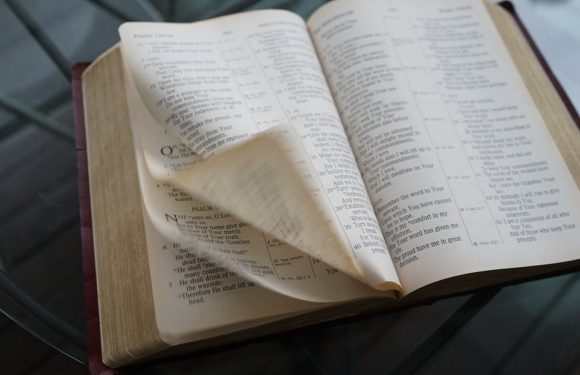An open Bible on a glass table, with pages inside that appear to be flipping or moving. The background shows a chair, suggesting a cozy reading space.