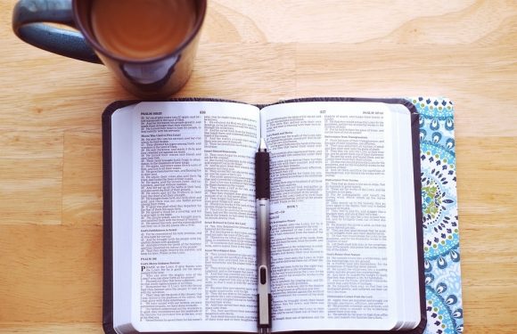 Open Bible on a wooden table with a floral-patterned cover, a black marker resting on it, and a mug of tea or coffee placed nearby.