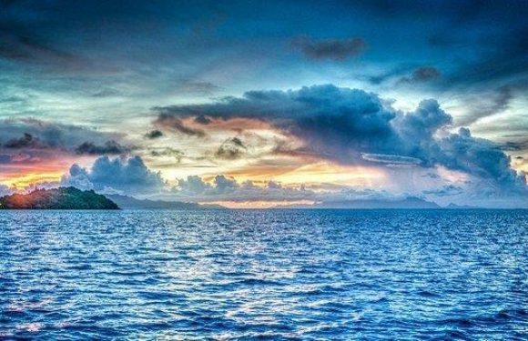 A coastal scene during sunset with a colorful sky, reflecting clouds, and an island in the distance; the water is calm with gentle waves.