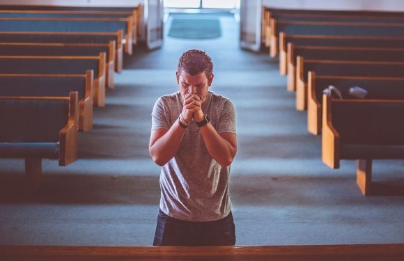 A young man stands in a church or chapel, with his hands clasped in front of his face, eyes closed, appearing to pray or reflect, surrounded by empty pews.