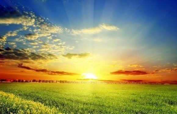 A vibrant sunset over an open green field with a partly cloudy sky and rays of sunlight streaming through the clouds.