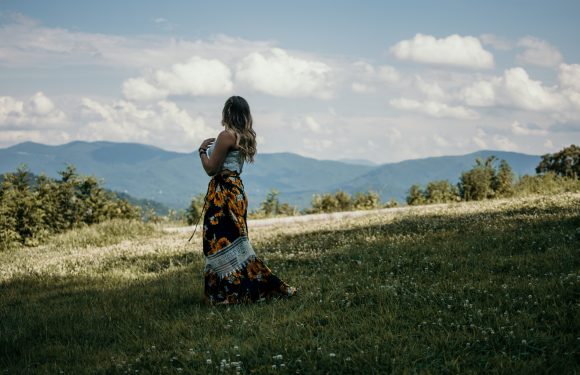 A woman in a floral dress stands in a grassy field with mountains and a partly cloudy sky in the background.