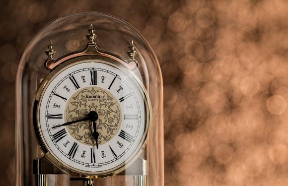 An elegant table clock with a glass dome, gold accents, and Roman numerals, showing the time as approximately 5:45, with a blurred warm-toned background.