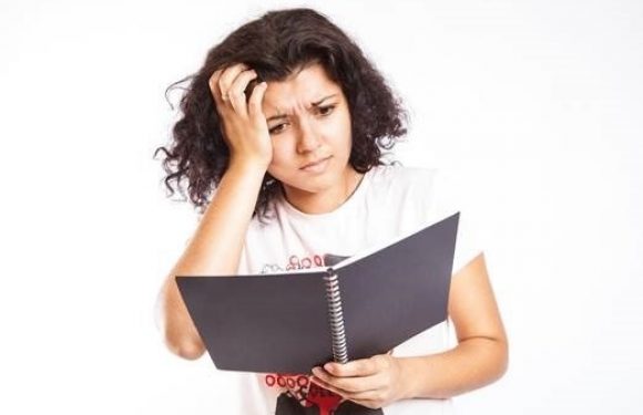 A woman with curly dark hair looking confused or puzzled while holding an open notebook and touching her head.