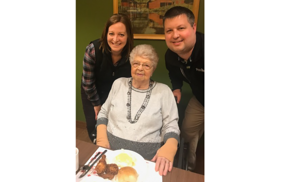 An elderly woman with white hair and glasses sitting at a table with a plate of food, flanked by a woman and a man standing beside her, all smiling.