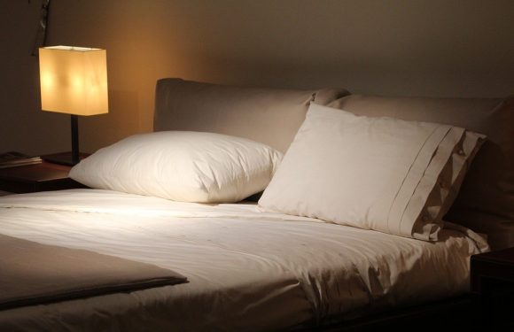 A neatly made bed with white sheets, multiple pillows, and a side table with a lit lamp in a softly lit bedroom.