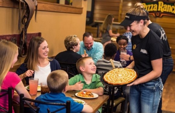 A smiling waitress in a black Pizza Ranch uniform serves a large apple pie to children and families dining at a restaurant with wooden interior decor and a cheerful atmosphere.