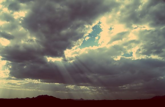 Cloudy sky with rays of sunlight breaking through, over a silhouette of distant mountains or hills.