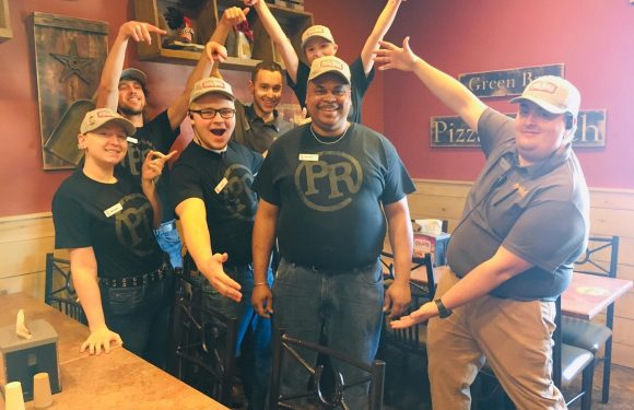 Group of seven people wearing Pizza Ranch uniforms and hats, smiling and posing enthusiastically inside a restaurant with red walls and wooden accents.