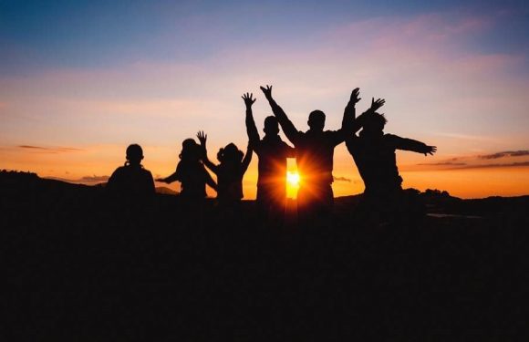 Silhouettes of six people with raised hands during sunset, standing outdoors on a hill under a colorful sky.