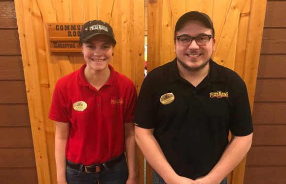 Two employees stand side by side in front of a wooden wall, smiling. They wear black caps, with the woman in a red shirt and the man in a black shirt, both with Pizza Ranch logos.