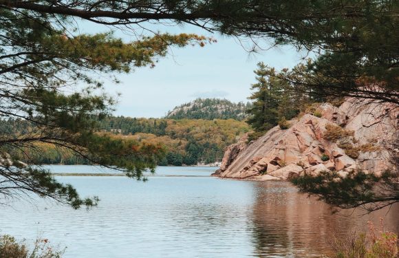 Two canoes rest on a rocky shoreline with trees framing a peaceful lake and distant hills in the background.