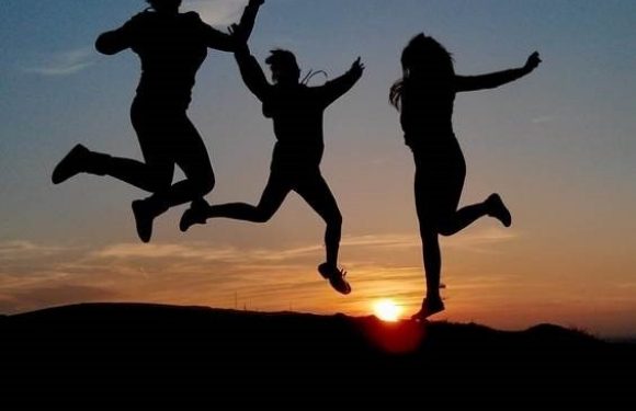 Three people in uniform are jumping and holding hands on a hill at sunset, silhouetted against an orange sky with a glowing sun near the horizon.