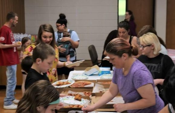 A group of people, including children and adults, gather around a table with pizza and food in a room with wooden accents, serving themselves from various dishes.