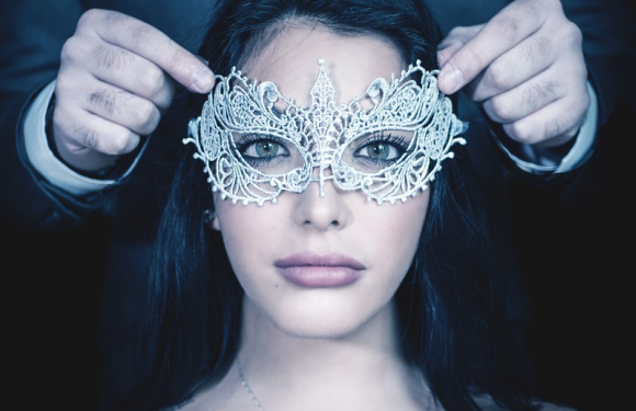 A woman with dark hair and fair skin wearing a decorative silver masquerade mask, while someone adjusts the mask on her face.