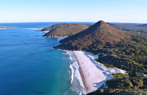 Aerial view of a coastline with two prominent hills covered in greenery, a sandy beach, and calm blue ocean waters extending into the horizon.