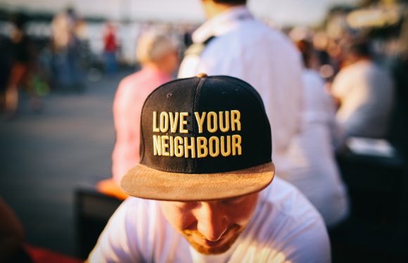 A person wearing a black baseball cap with yellow embroidered text reading "LOVE YOUR NEIGHBOUR" and a tan brim, outdoors with a blurred crowd in the background.