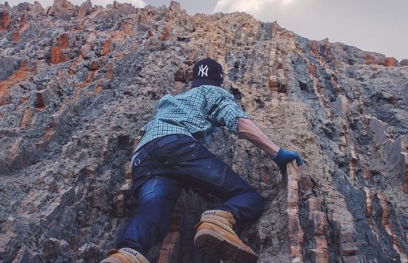 Person climbing rocky cliff face wearing a New York Yankees cap, blue gloves, plaid shirt, jeans, and tan work boots.