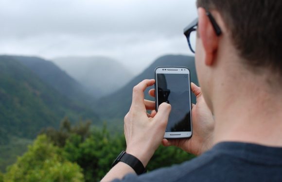 A person wearing glasses and a black shirt takes a photo with a smartphone of a mountainous landscape with green trees and misty clouds.