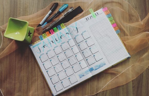 A January calendar page on a grid notebook with colorful tabs, black pens, a green star-patterned bowl, and sheer yellow fabric on a wooden surface.