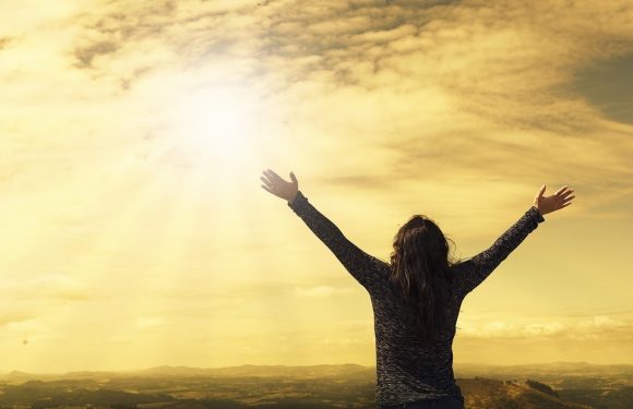 A woman stands with arms raised outdoors during sunset, with a partly cloudy sky and a distant landscape in the background. She is wearing a dark long-sleeve shirt.