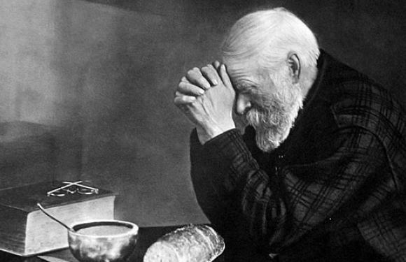 An elderly man with a white beard and hair, sitting with his head bowed and hands covering his face, in front of a loaf of bread, a bowl, and a small box.
