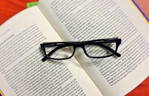 A pair of black-rimmed eyeglasses resting on an open book with text, on a surface with a hint of orange and green background.