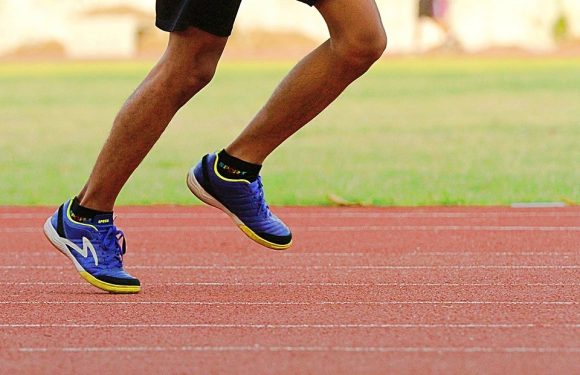 A person running on a track, wearing black shorts, blue running shoes with yellow accents, and black ankle socks.