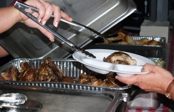 Person serving cooked meat onto a plate at a buffet with tongs, using a white plate and a chafing dish filled with meat.