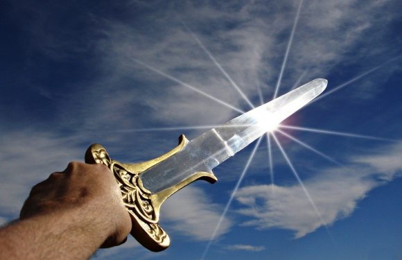 A hand holds a golden ornate dagger with a clear blade pointed towards the bright sun in a partly cloudy sky.