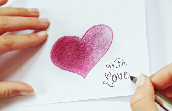 A person is writing "With Love" in black ink below a pink and red heart drawn with colored pencils on white paper.