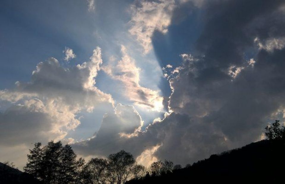 Clouds partially cover the sun in a sky with light filtering through, above a silhouette of trees and a hill.