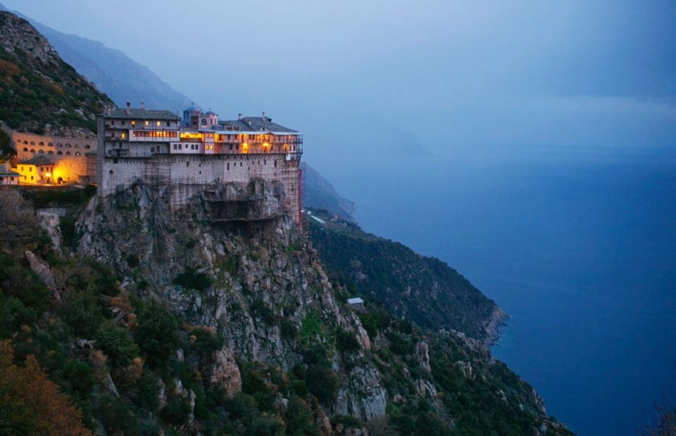 A large building sits atop a steep cliff overlooking the ocean, illuminated by warm yellow lights, with a mountain landscape in the background during dusk.