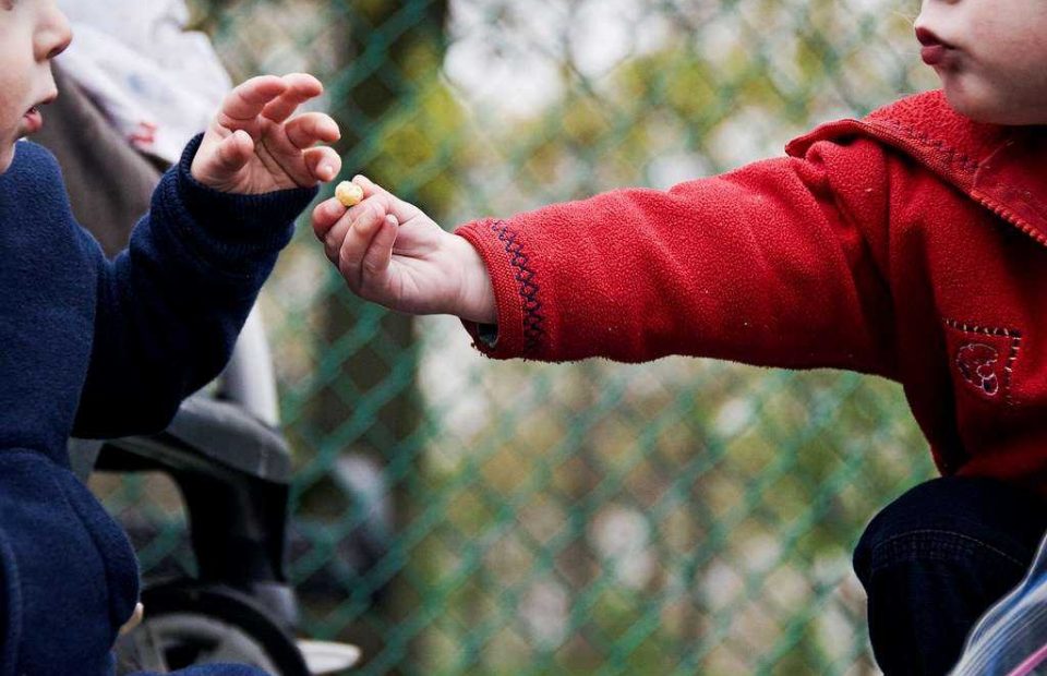 Two children exchanging a small snack outdoors, one wearing a red jacket and the other a navy blue jacket, behind a green chain-link fence.