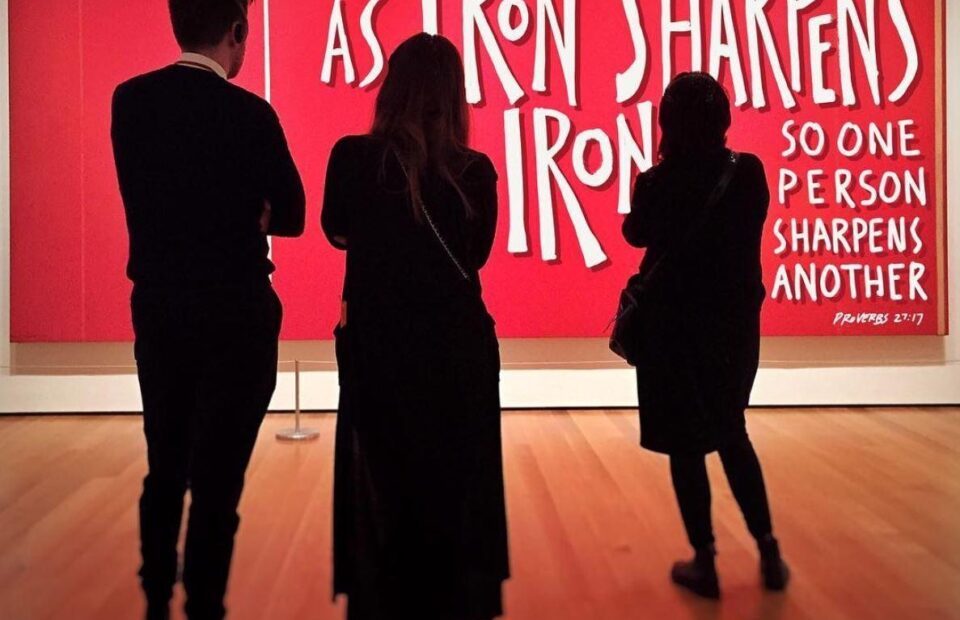 Three people stand in front of a red background with text, observing the artwork. One person wears a black dress, and the others are dressed in dark clothing.