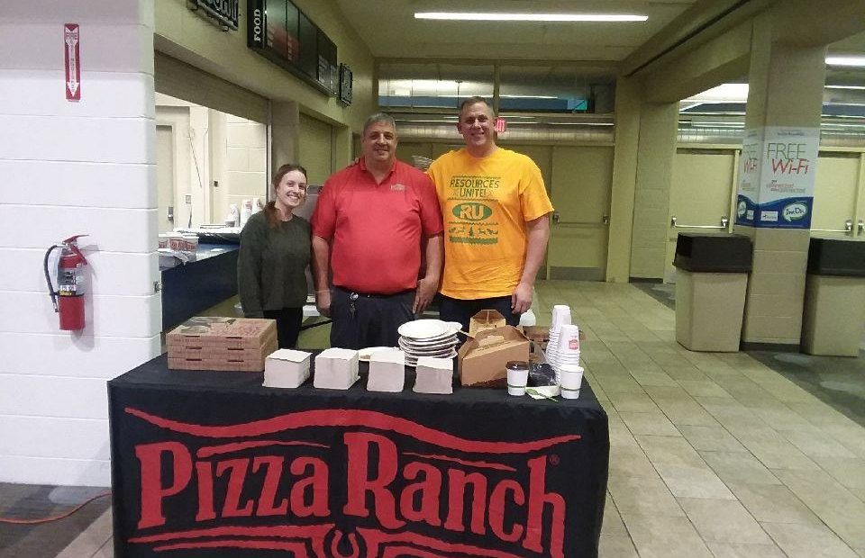 Three people standing behind a table with pizza boxes, cups, and takeout containers, in front of a Pizza Ranch tablecloth, in an indoor hallway.