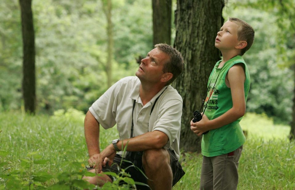 A man and a boy are in a lush green forest, both looking upward with curious expressions. The man is kneeling, wearing a light-colored polo shirt, and the boy is standing in a green sleeveless shirt with a lanyard and binoculars.